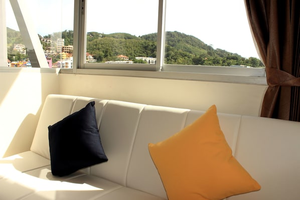 A white sofa with two pillows, one dark and one light, in a room with a large window overlooking a green hillside and buildings.