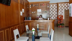 A hotel lobby with a wooden reception desk, a glass-top table with chairs, a ceiling fan, and a decorative wall with a geometric pattern. - Tropical Sunset Hotel Patong (Patong)