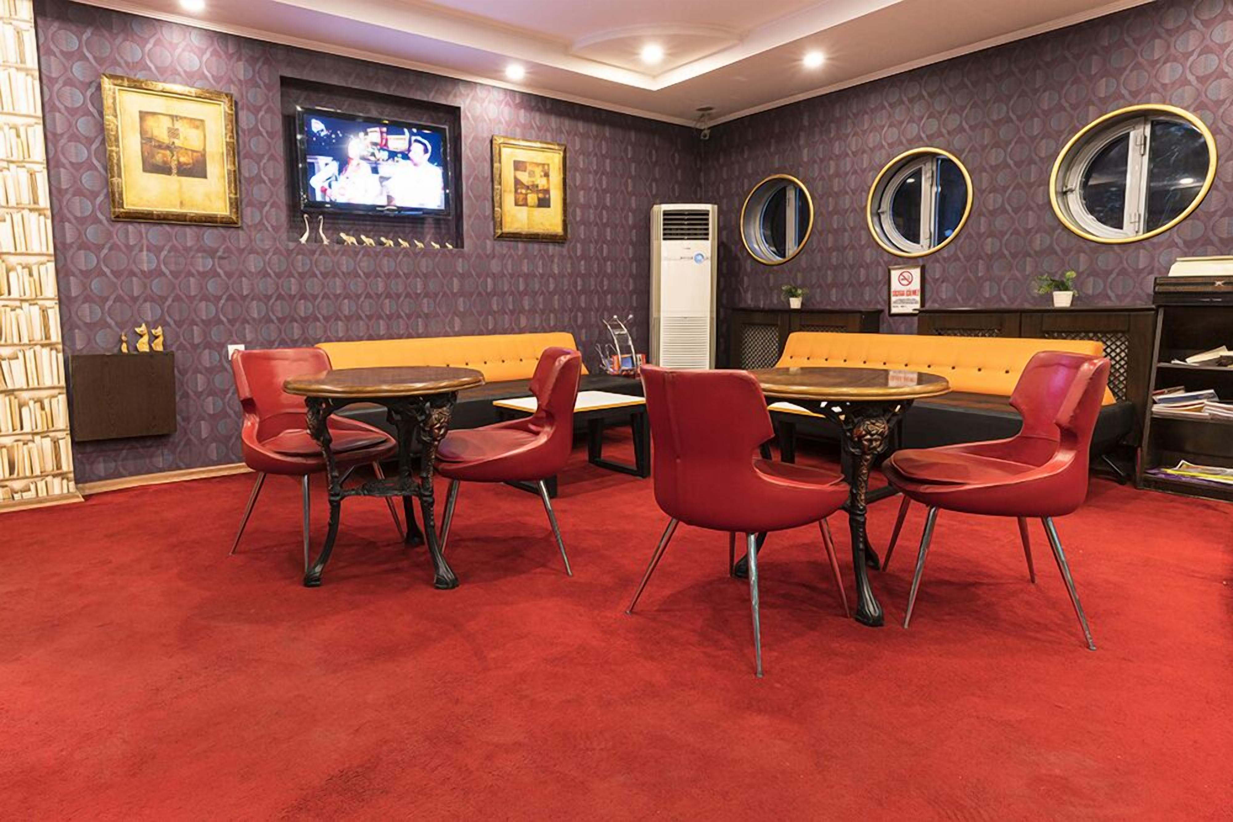 A room with a red carpet, a sofa, a table, and chairs. There is a television on the wall and a sign with a prohibition symbol.