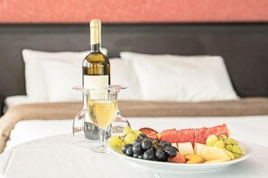 A bed with white linens, a bottle of white wine, a glass pitcher, and a plate of assorted fruits.