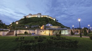 Un castillo histórico en una colina con un edificio moderno en primer plano, rodeado de jardines cuidados y farolas.