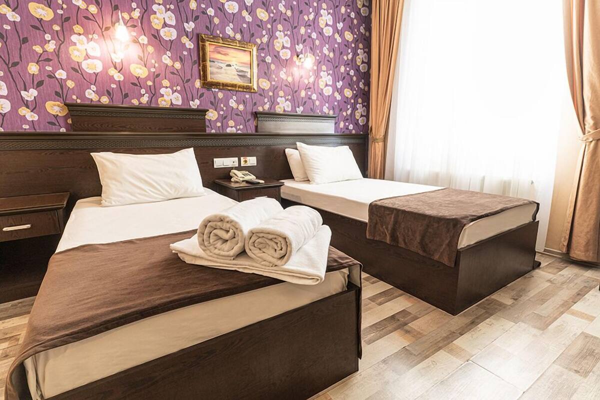 Een hotelkamer met twee bedden, een houten hoofdeinde, bloemenbehang en een raam met gordijnen.