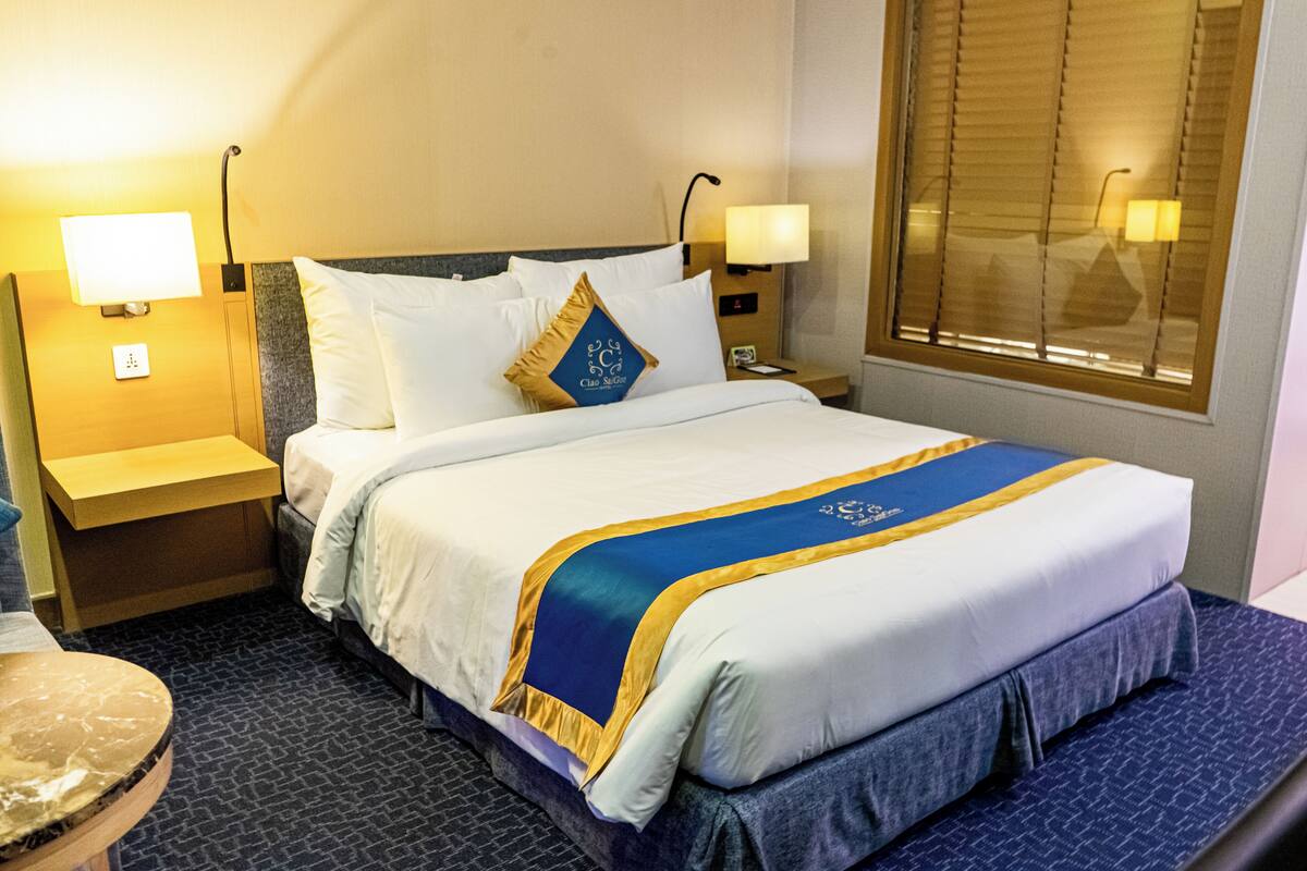 Quarto de hotel com uma cama bem arrumada, duas mesas de cabeceira com abajures, uma janela com persianas e piso coberto por carpete.