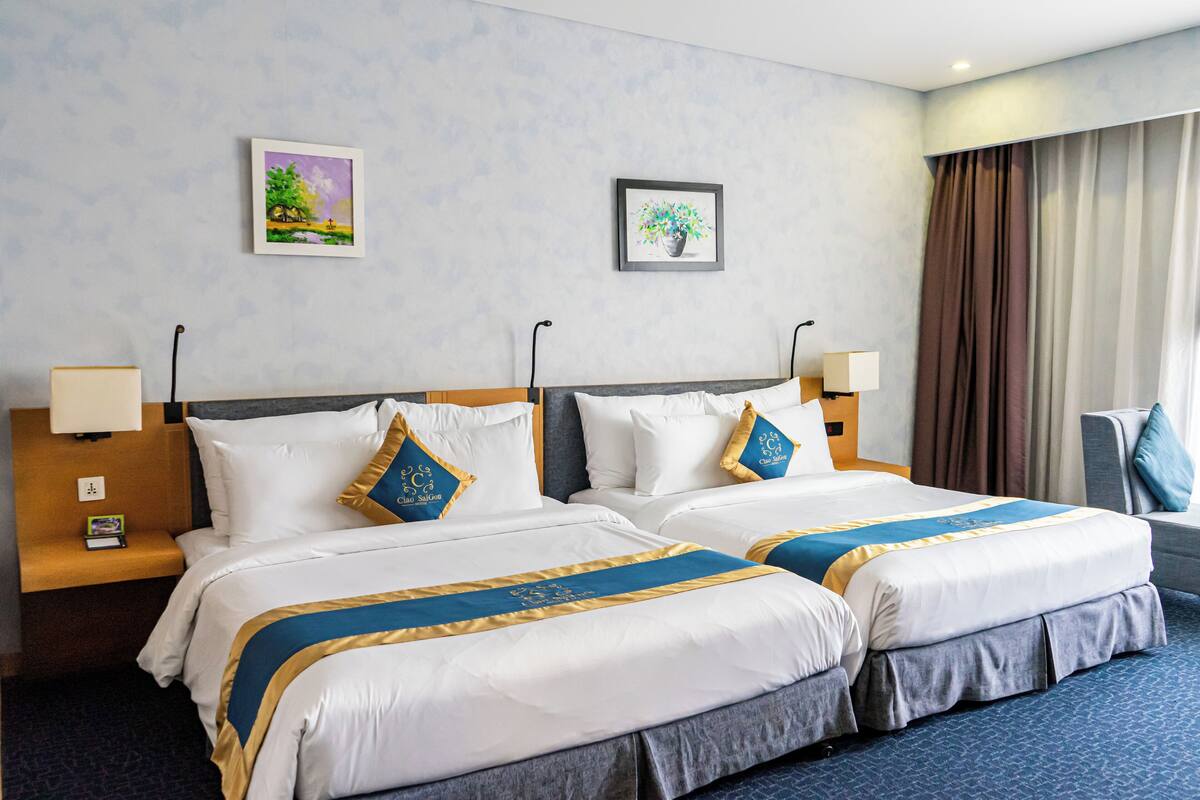 Uma cama de casal com roupa de cama branca e azul, duas mesas de cabeceira com abajures e duas fotos emolduradas na parede.