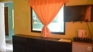 Una habitación con ventana cubierta por una cortina, un escritorio con computadora, un refrigerador pequeño y un televisor en la pared.