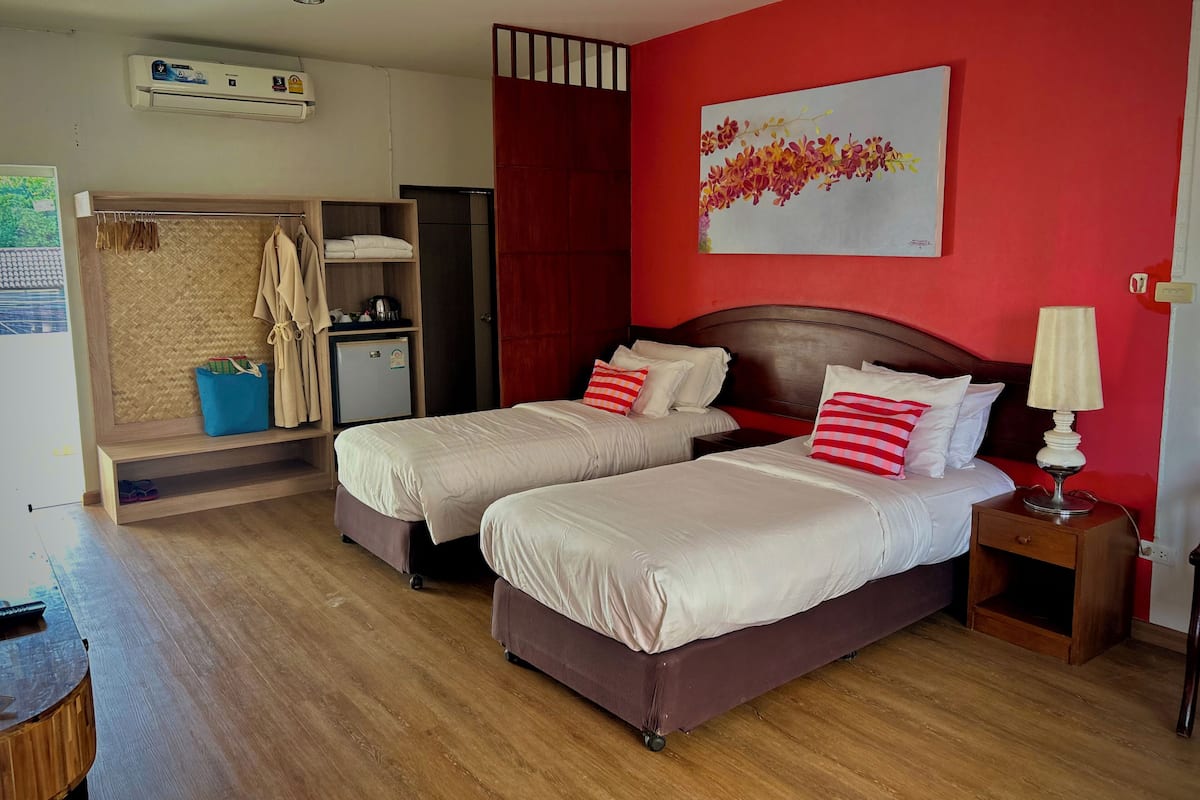 Una habitación con dos camas, un escritorio pequeño, un armario, una pared roja con un cuadro floral y un ventilador de techo.