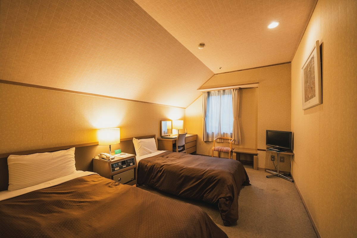 A hotel room with two beds, a desk, a chair, a TV, and a window with curtains.