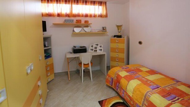 Une chambre à coucher de taille modeste, comprenant un lit simple, un bureau et une chaise. On y trouve une armoire avec plusieurs tiroirs et une étagère au-dessus du bureau.