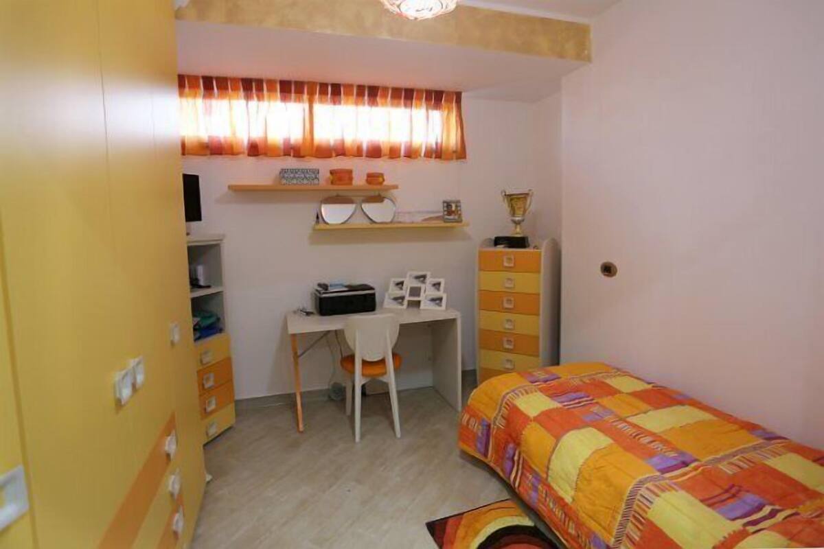 Una habitación pequeña con cama individual, escritorio y silla. Se observa un armario con varios cajones y un estante sobre el escritorio.