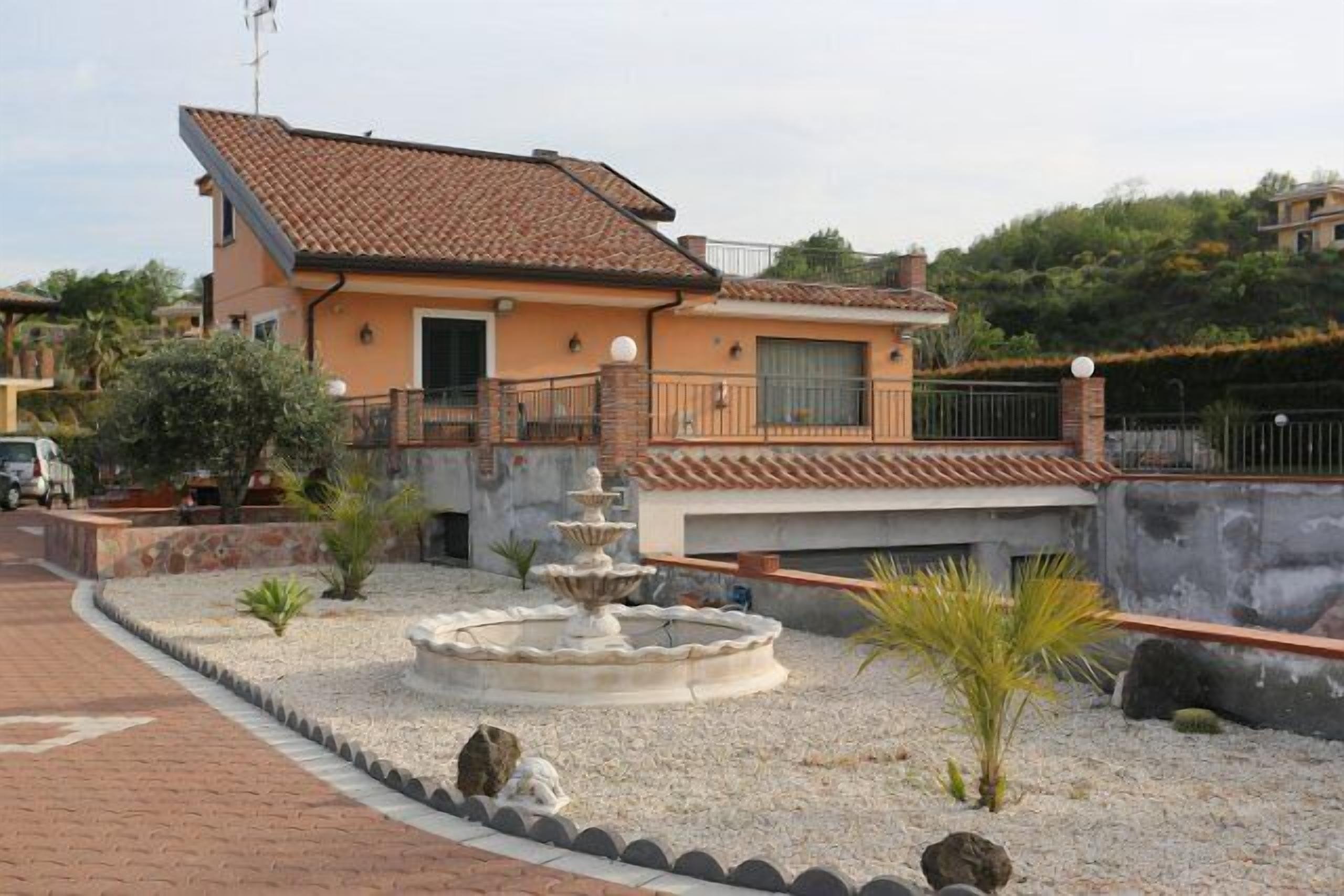 A two-story house with a tiled roof, a small garden with a fountain, and a paved driveway.