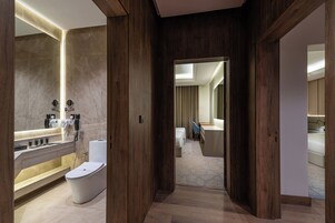 A modern bathroom with a bathtub, toilet, and vanity. There is a wooden floor and a view into a bedroom with a bed and a desk.