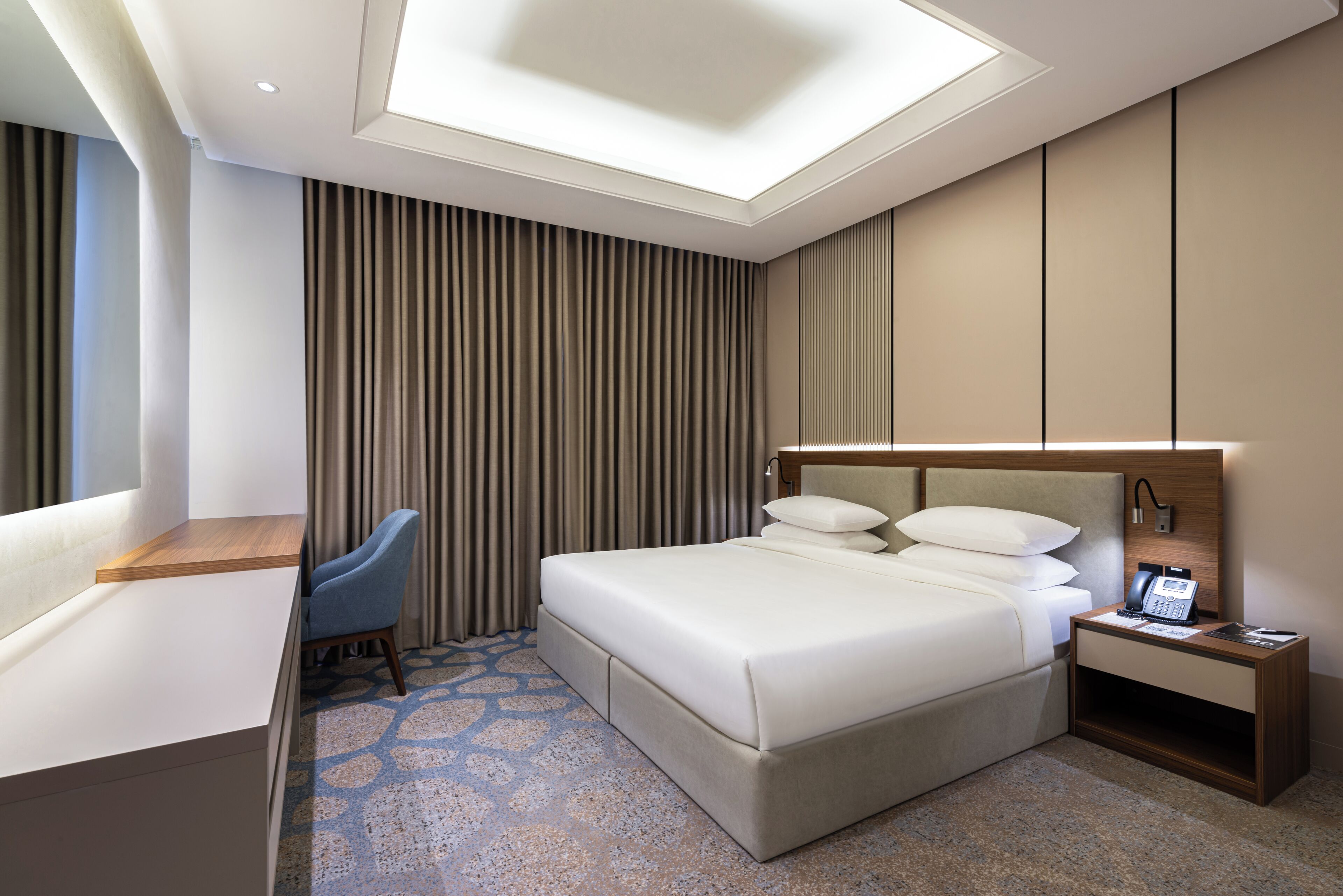 A modern hotel room with a large bed, a desk, a chair, and a nightstand with a phone.