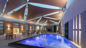 Una piscina con iluminación azul en su interior, rodeada de mesas y sillas, bajo grandes estructuras de sombra triangulares.