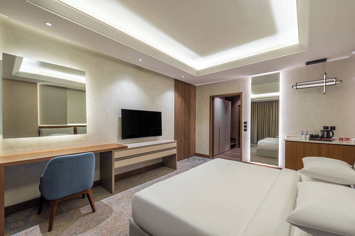 Una habitación de hotel moderna con una cama grande, un escritorio con una silla, un televisor, un espejo y una pequeña cocina.