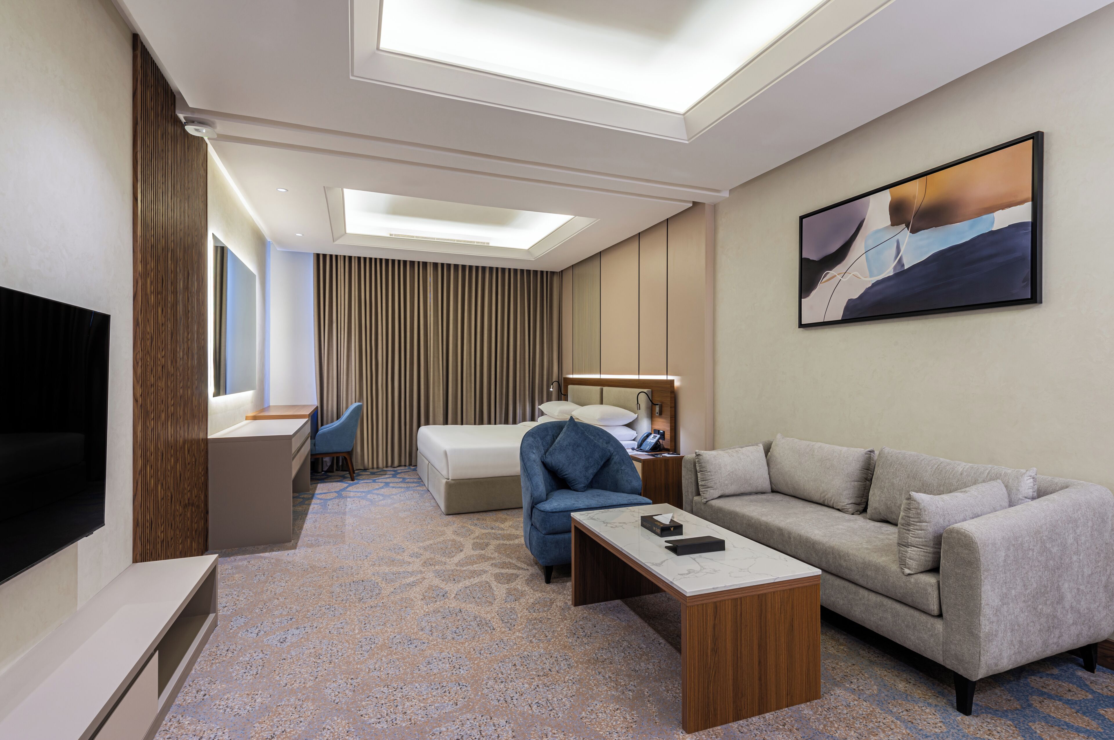 A hotel room with a bed, a sofa, a desk, a chair, a TV, and a painting on the wall.