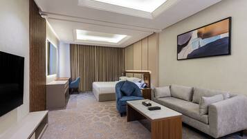 A hotel room with a bed, a sofa, a desk, a chair, a TV, and a painting on the wall.