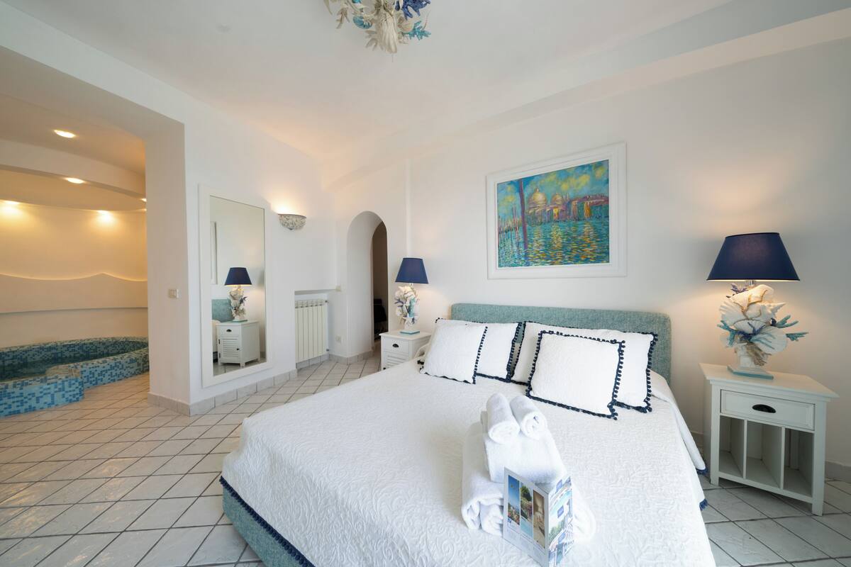 A neatly arranged bedroom with a large bed, a painting on the wall, and a small table with a lamp.