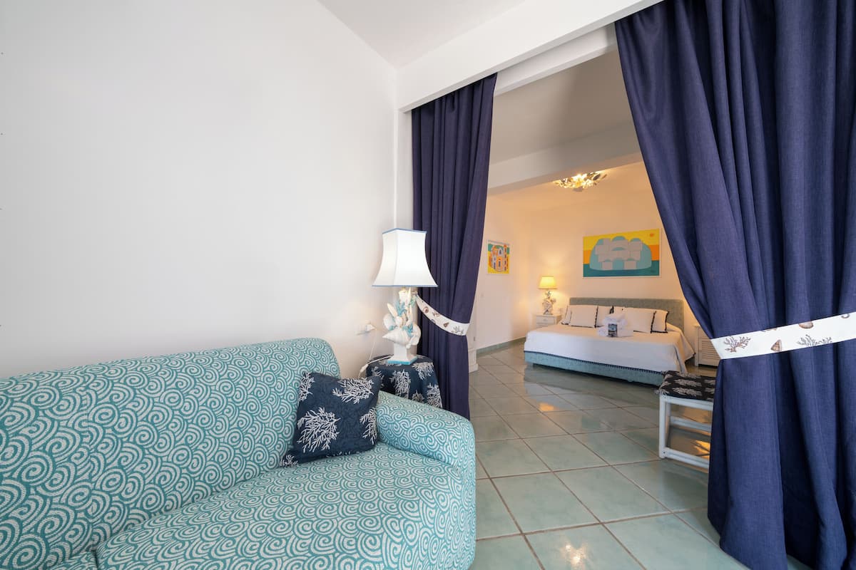 A room with a teal patterned sofa, a white lamp, and a bed with a white and blue bedspread.