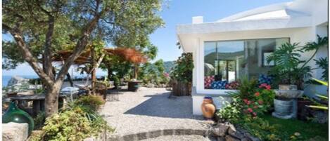 Une maison moderne avec un chemin de gravier menant à une porte en verre, entourée d’une végétation luxuriante et offrant une vue sur la mer.