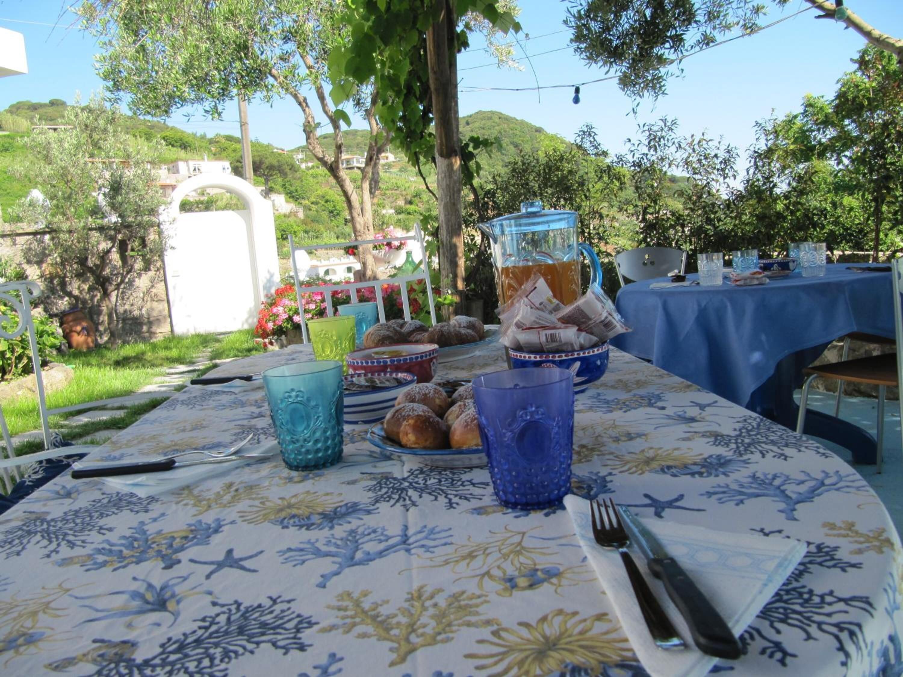 Una mesa para comer al aire libre con mantel floreado, rodeada de follaje y árboles.