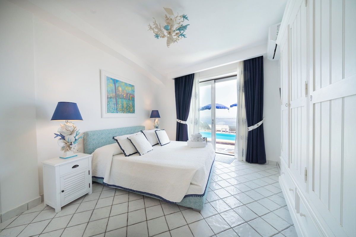 A bedroom with a bed, two bedside tables, a painting, and a view of the pool.