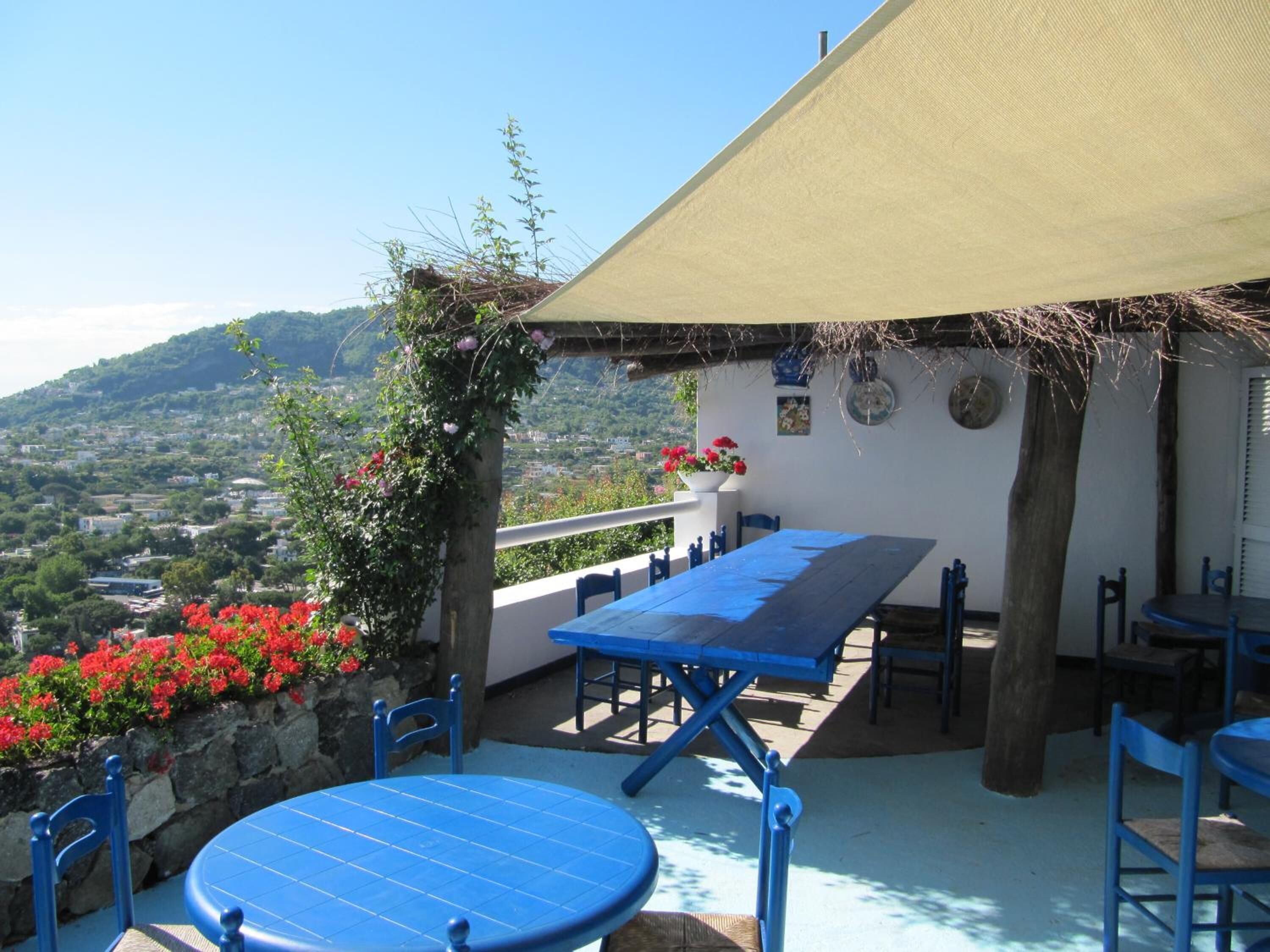 Un patio al aire libre con mesa y sillas, techo de paja y vista a un pueblo en la ladera.
