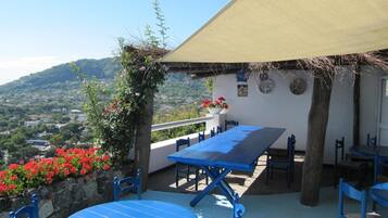 Un patio al aire libre con mesa y sillas, techo de paja y vista a un pueblo en la ladera.