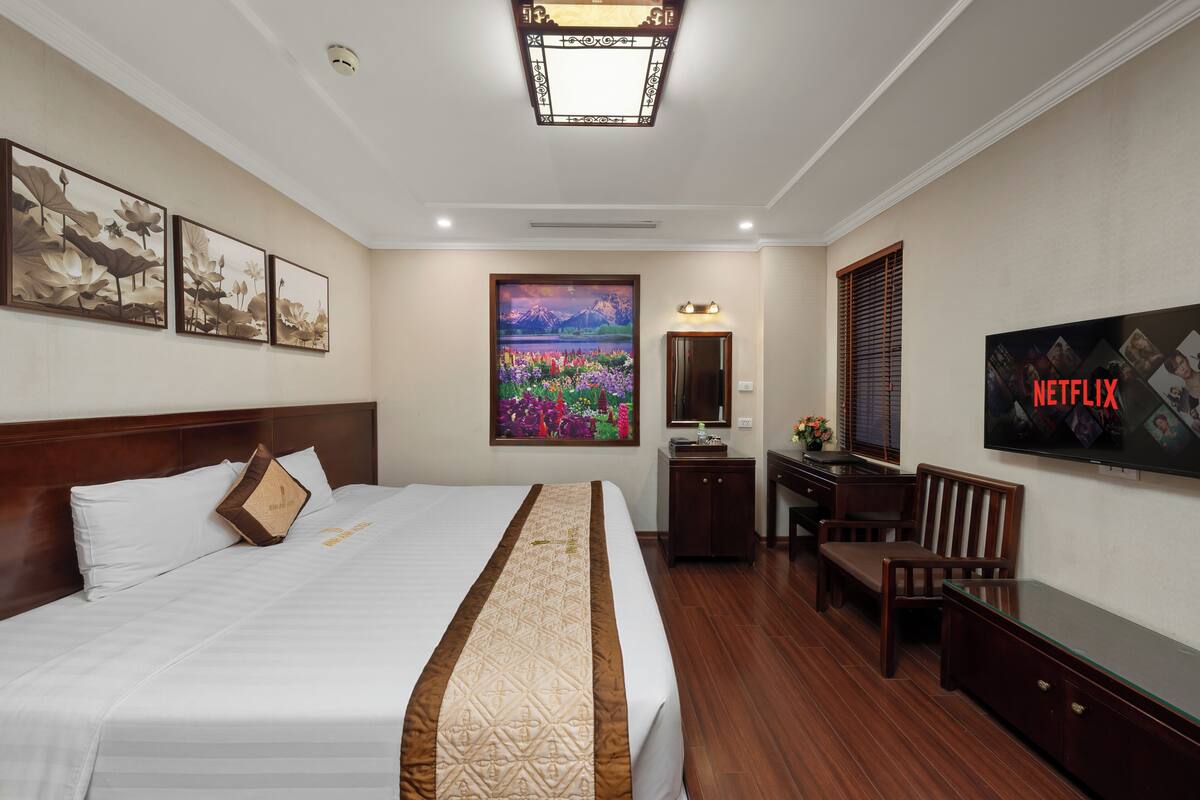A hotel room with a large bed, a desk, a chair, a TV, and artwork on the walls.