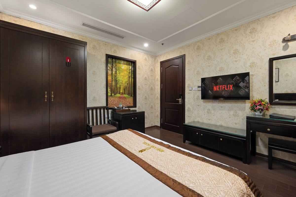 A hotel room with a bed, wardrobe, desk, TV, and a painting of a forest.
