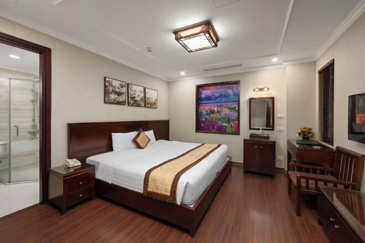 A hotel room with a large bed, bedside tables, a desk, a chair, a painting, a mirror, and a television.
