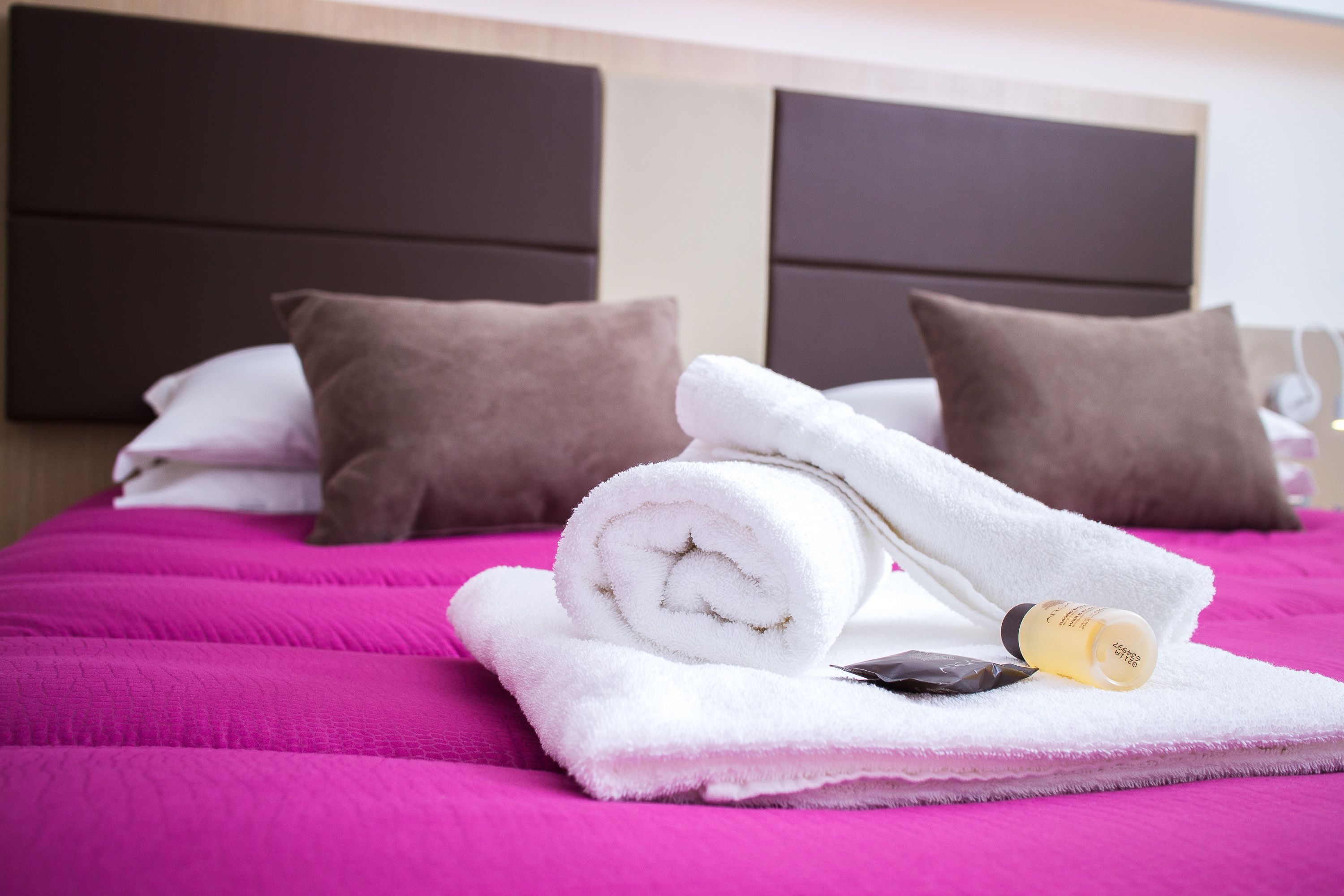 Ein ordentlich bezogenes Bett mit einer rosafarbenen Tagesdecke, weißen und braunen Kissen sowie einem aufgerollten weißen Handtuch und einer Lotionflasche.