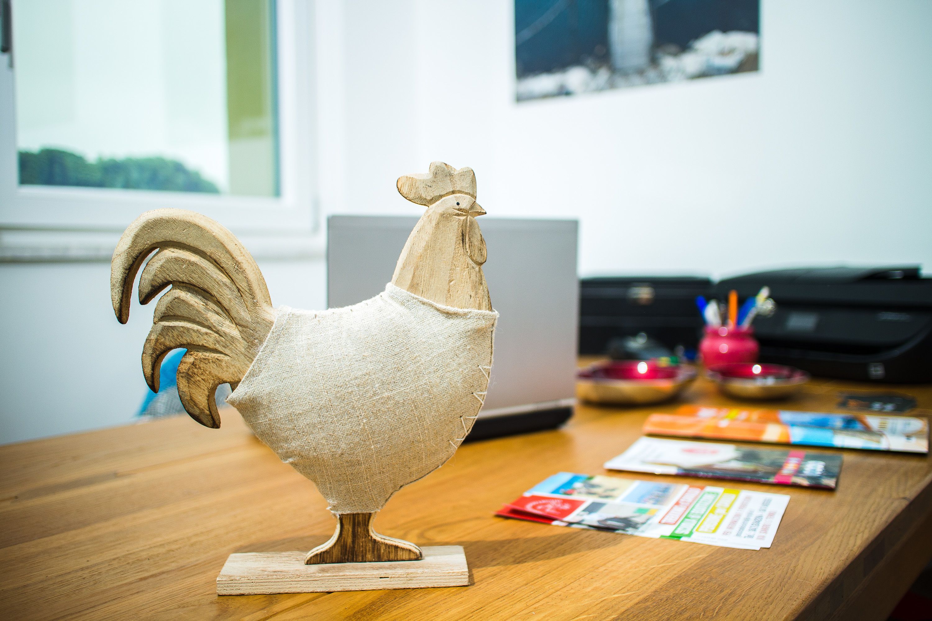 A wooden rooster sculpture on a wooden table with a laptop and some brochures in the background.