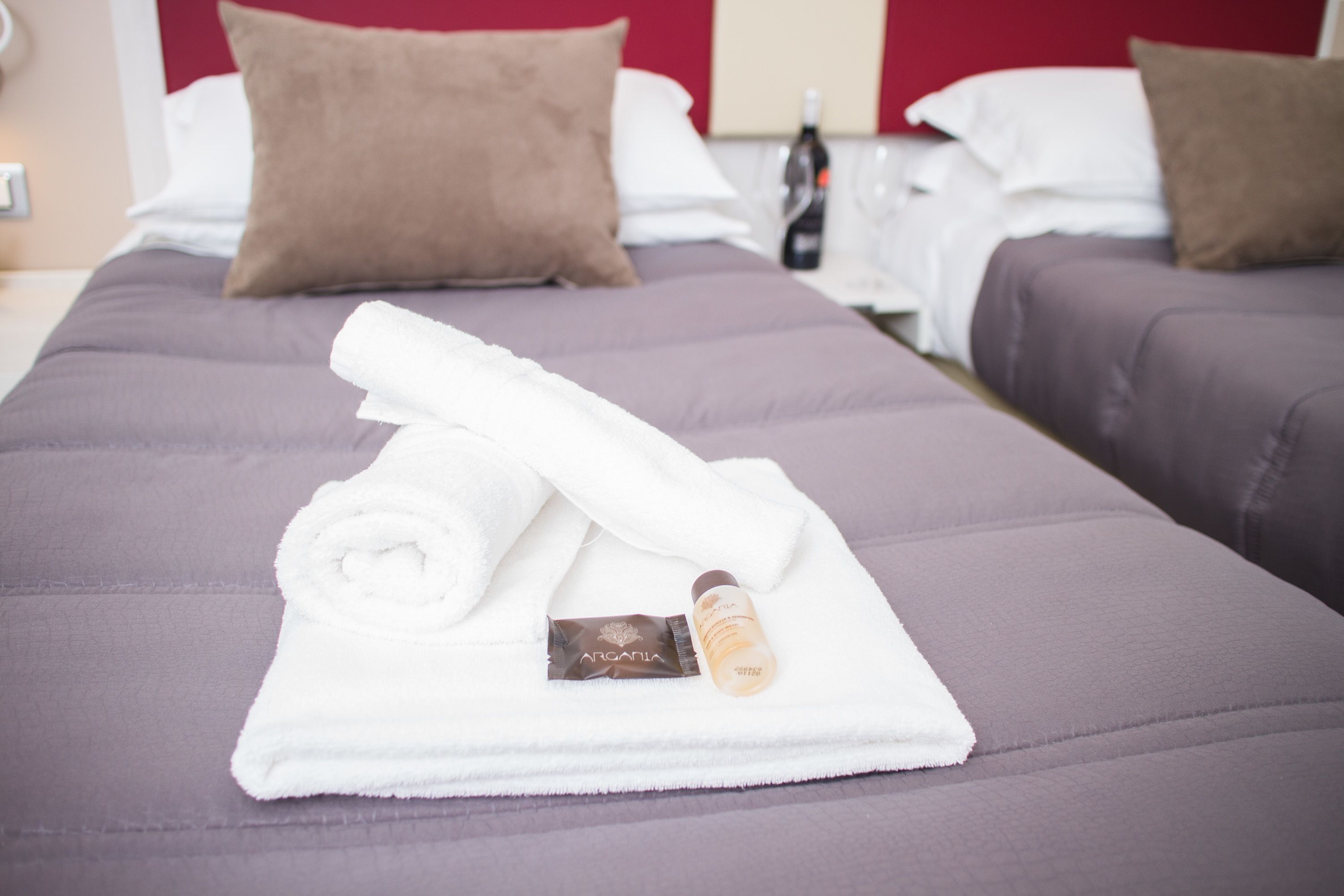 A neatly made bed with a white towel, a bottle of lotion, and a small card on a purple bedspread in a hotel room.