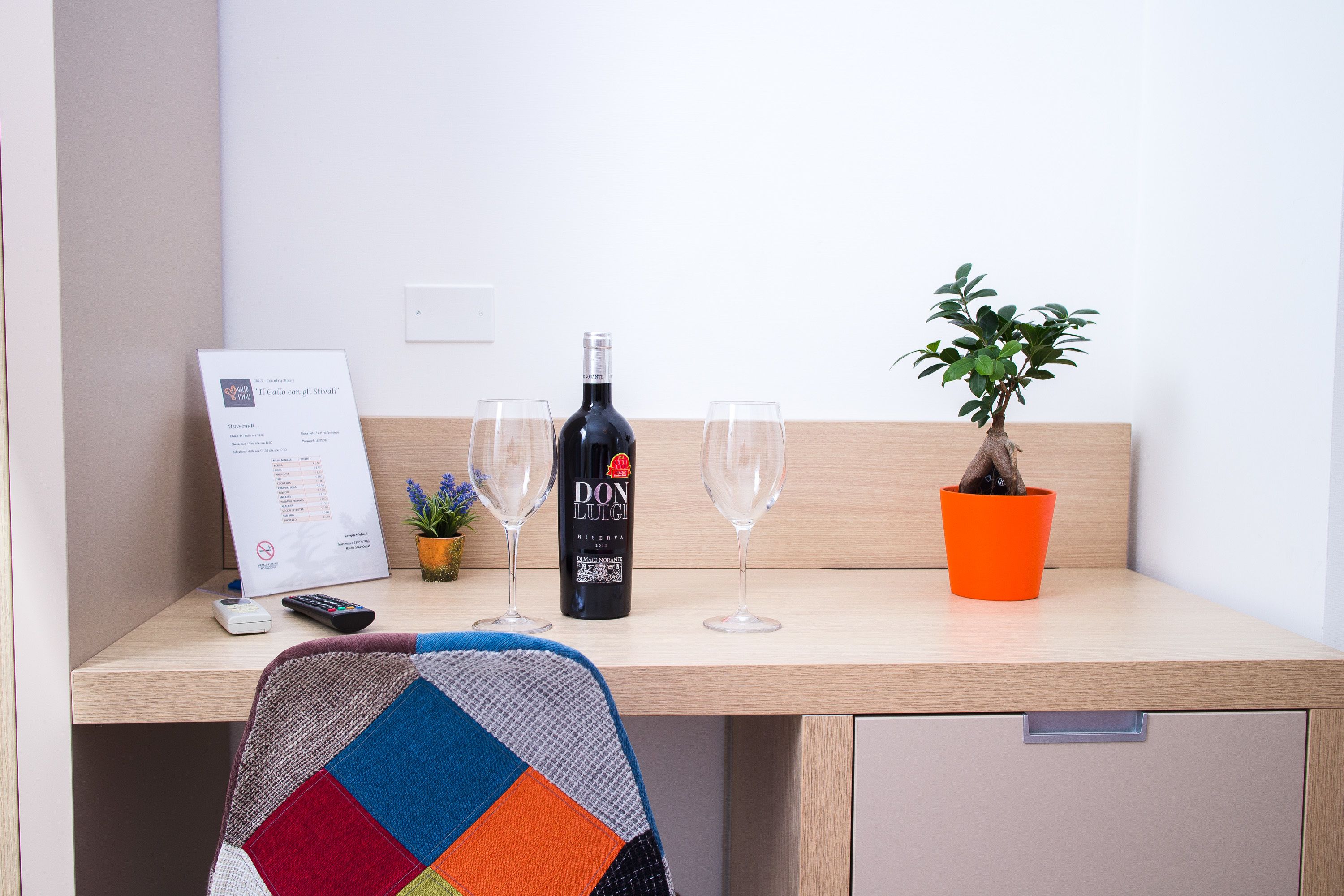 A hotel room with a wooden desk, a bottle of Don Luigi wine, two glasses, a remote control, a small plant, and a colorful chair.