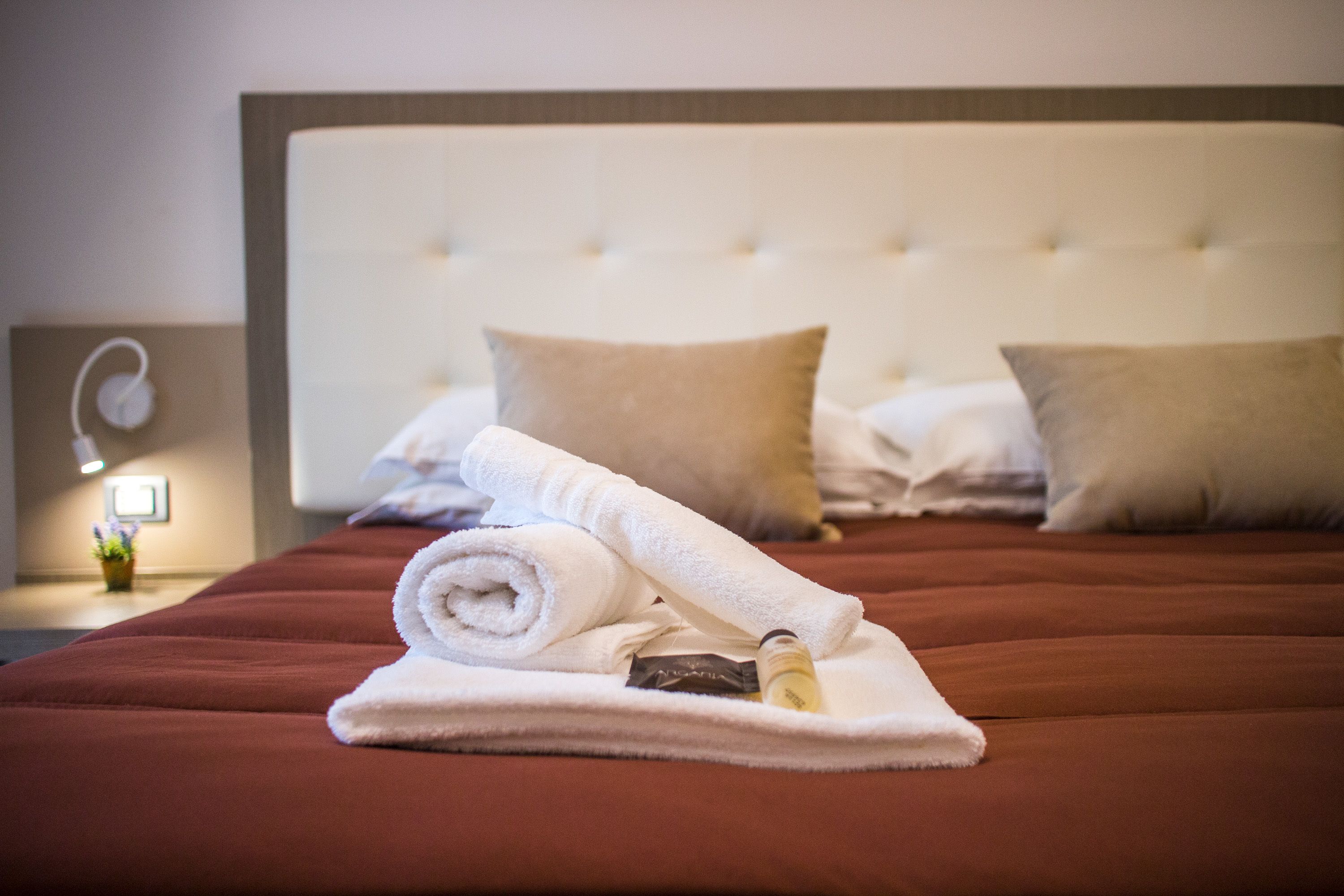 A neatly made bed with a rolled white towel and a bottle on it, a white headboard, and two pillows.