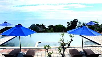 A poolside area with blue umbrellas, lounge chairs, and a view of the sea.