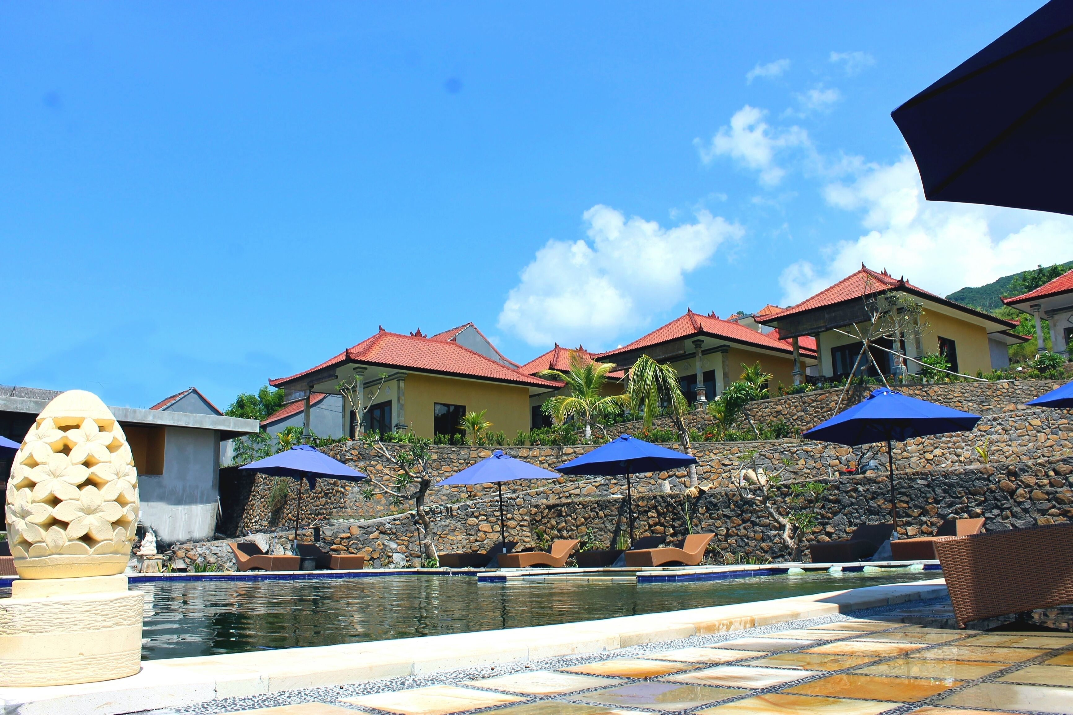 A pool area with lounge chairs and umbrellas, a stone wall, and buildings with red roofs.
