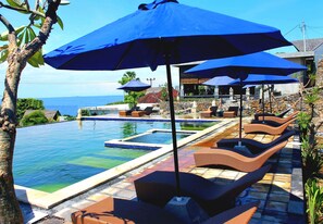 Zona de piscina con sillones reclinables y sombrillas azules, una pared de piedra y vista al océano.
