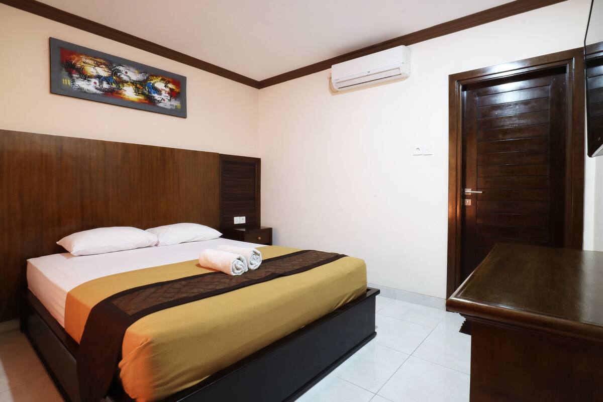 A hotel room with a bed, a nightstand, a wooden headboard, a painting on the wall, and a wardrobe.