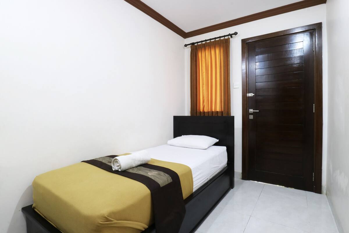A single bed with a brown headboard, a white pillow, and a yellow and black bedspread. There is a dark wooden door with a brown curtain to the right.