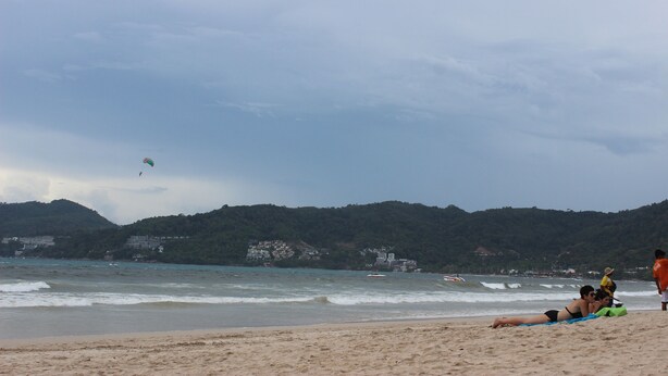 A beach with people relaxing, a kite in the sky, and buildings in the background. - Tulip Inn (Patong)