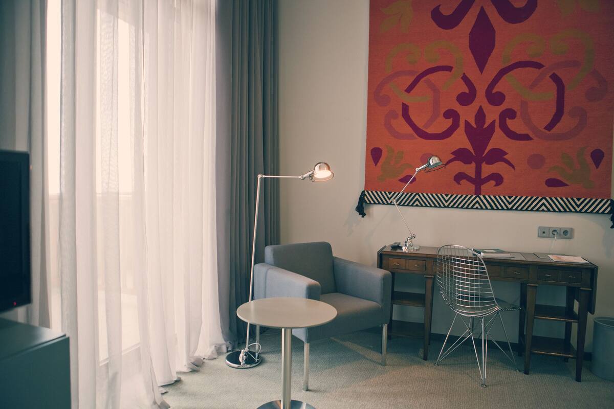 A hotel room with a desk, chair, and a large window with sheer curtains.