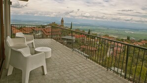 Ein Balkon mit zwei weißen Plastikstühlen und einem kleinen runden Tisch, mit Blick auf eine Stadt mit einem markanten Turm und grünen Feldern.