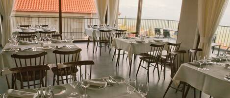Un restaurante con varias mesas dispuestas para comer, ventanas amplias con cortinas y vista a un edificio de tejas rojas.