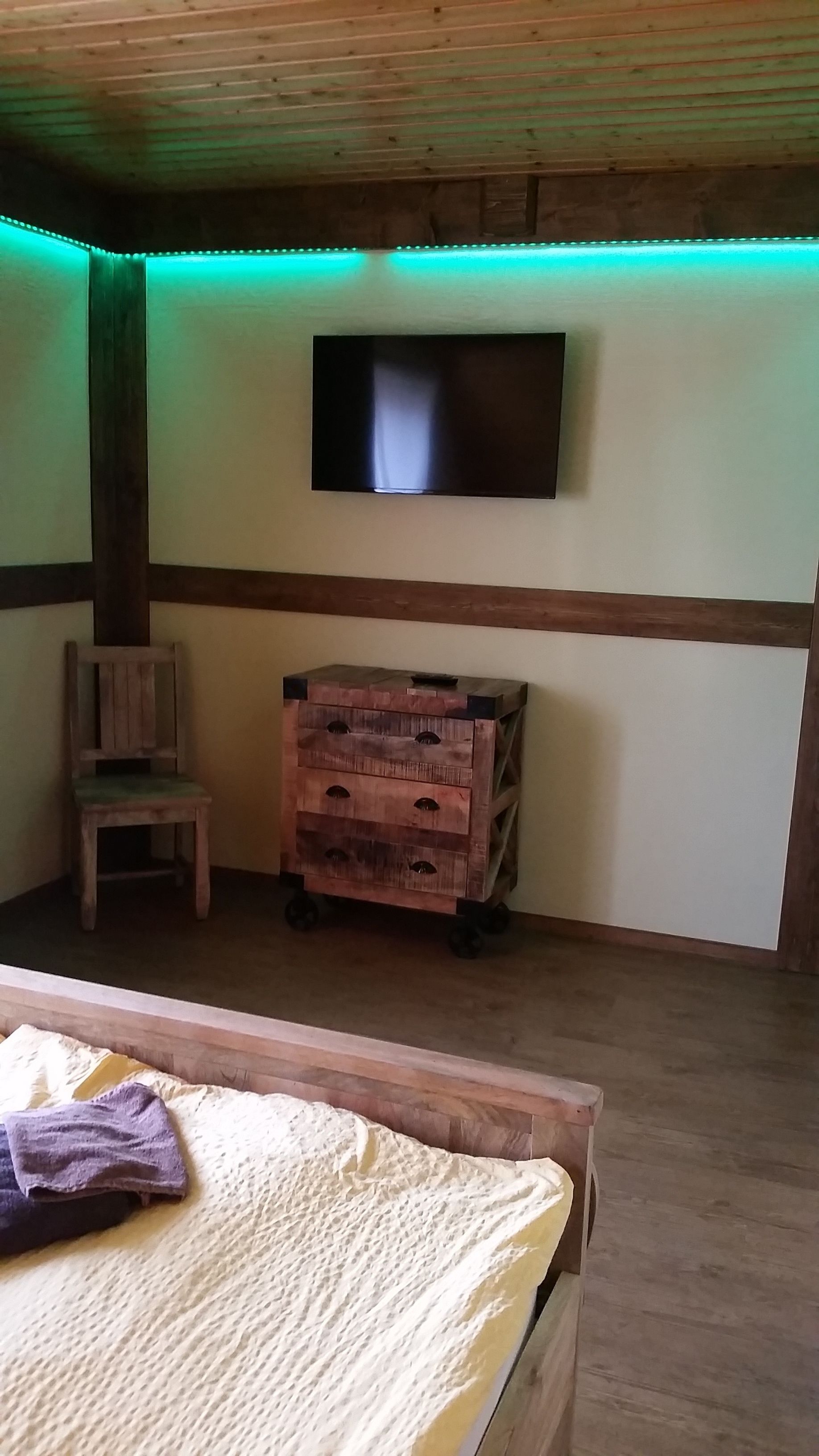 Une chambre à coucher comprenant un lit, une chaise en bois, une petite commode en bois et un téléviseur fixé au mur.