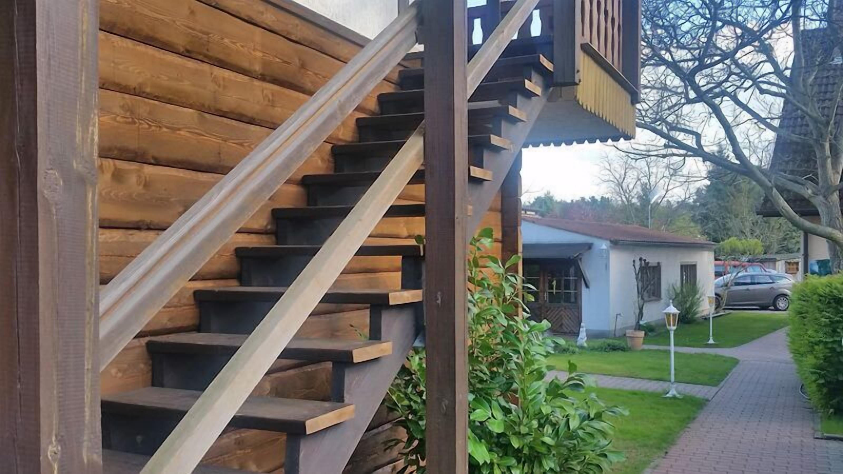 Eine Holztreppe mit Metallgeländer, die zu einem Gebäude mit weißer Fassade und einem kleinen Garten führt.
