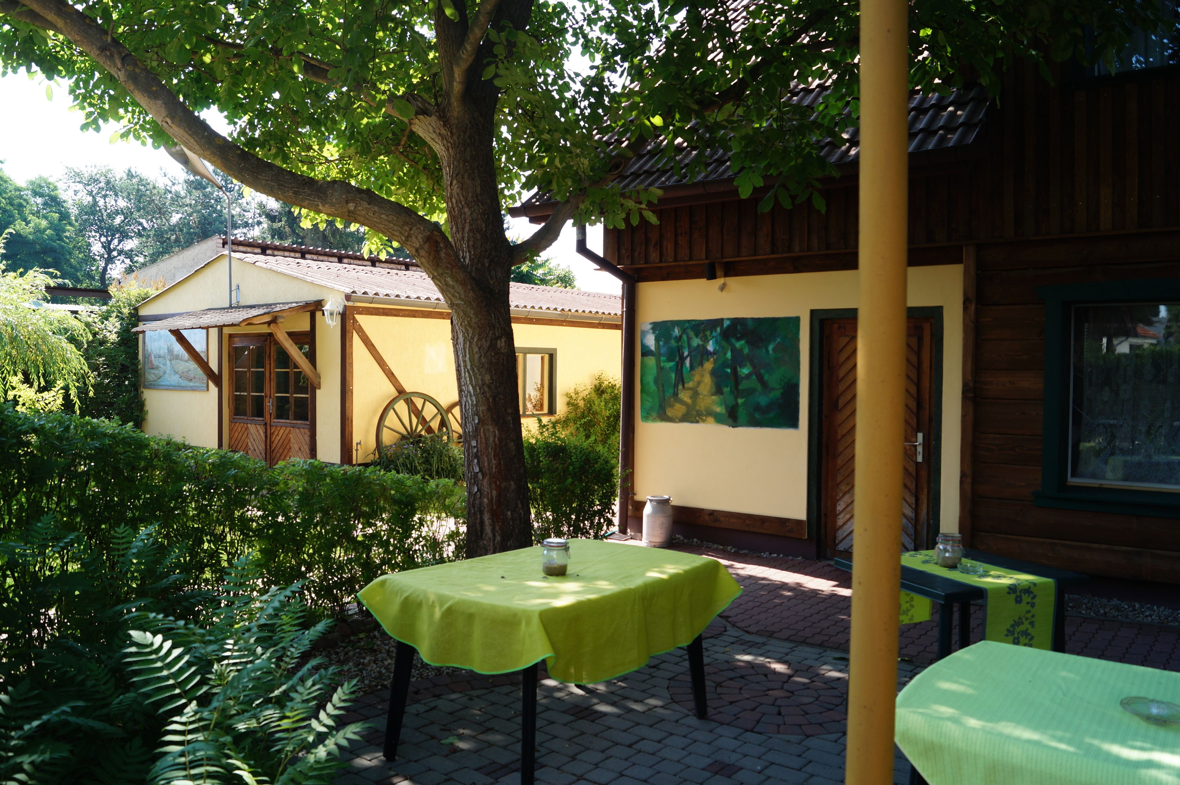 A courtyard with a table covered in a green tablecloth, a wooden table, and a chair. There is a tree providing shade, and a building with a window and a door in the background.