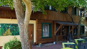 Un edificio rústico de madera con una puerta verde, una ventana y una escalera que conduce a un nivel superior. Hay mesas y sillas al aire libre, y un árbol en primer plano.