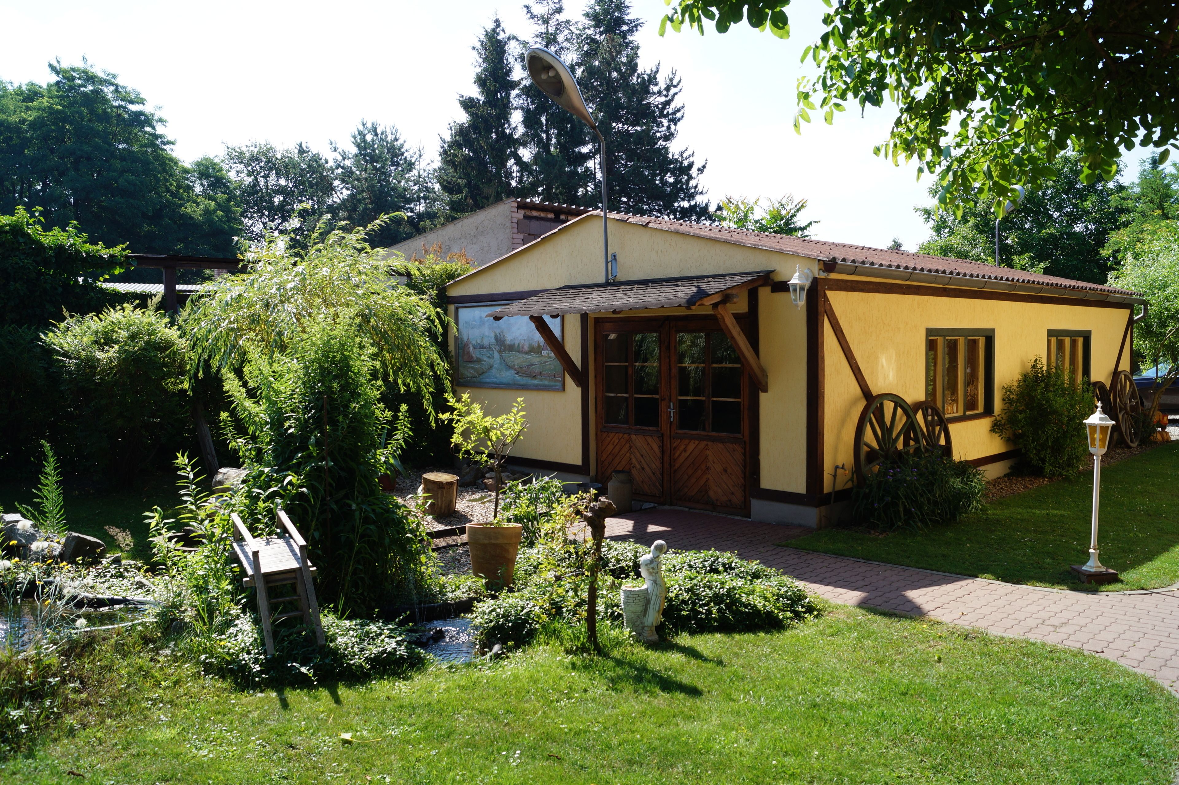 Un petit bâtiment jaune, doté d’une porte et de fenêtres en bois, entouré de verdure et d’un petit étang.