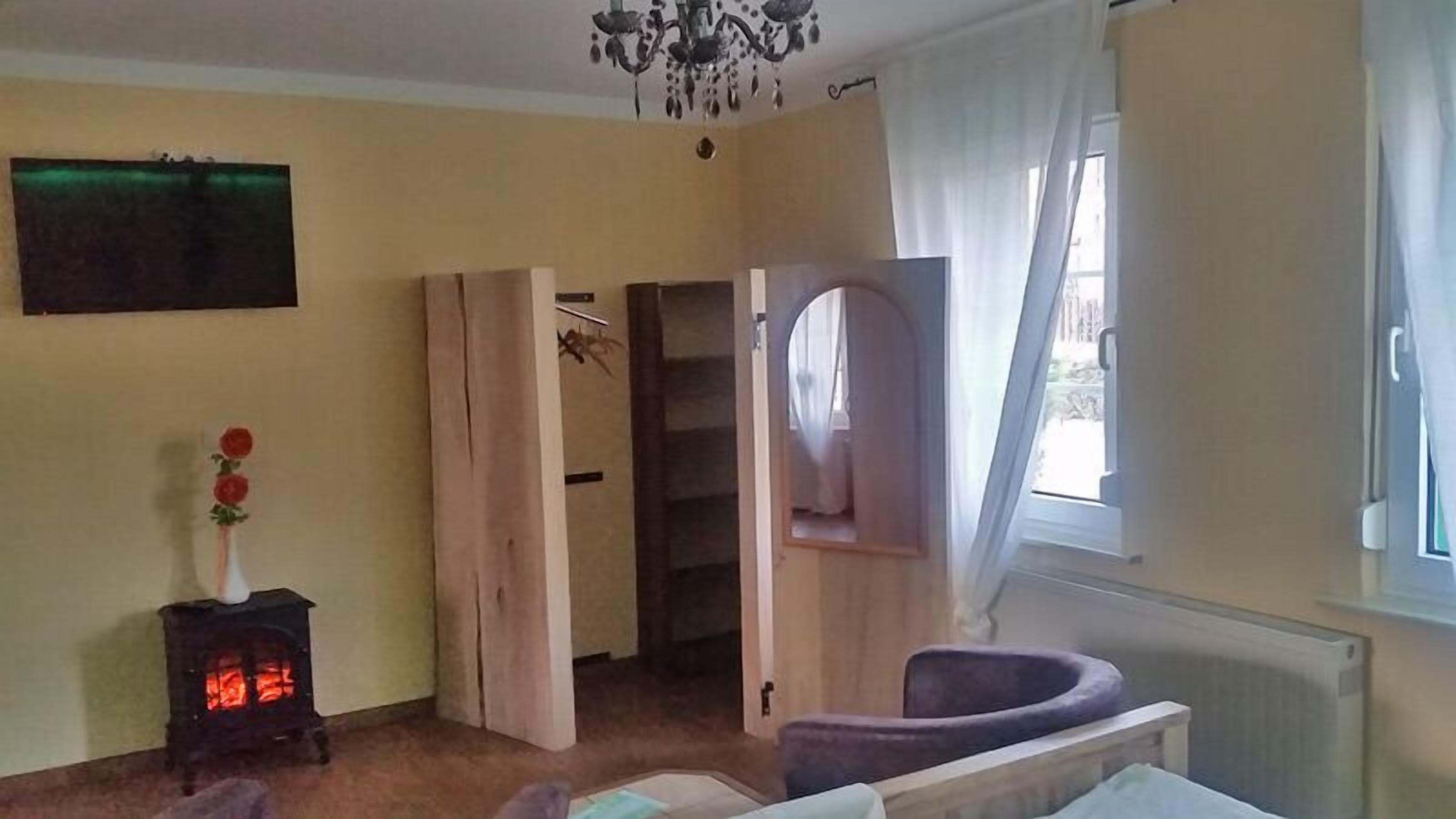 Quarto com porta de madeira que dá para uma escada, um pequeno aquecedor com chama vermelha, uma cadeira e uma janela com cortinas.